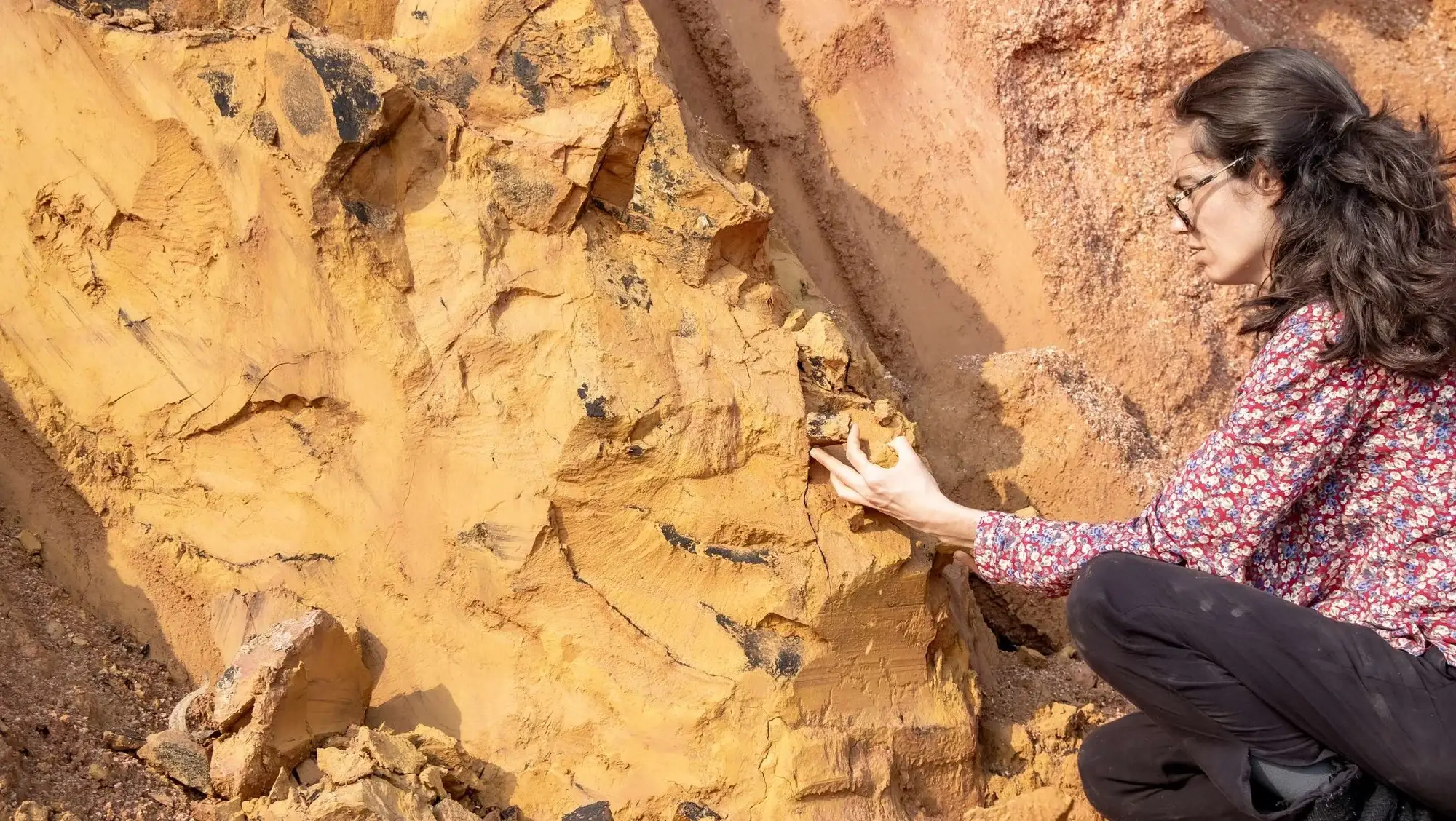Marcela da Terra agachada diante de um barranco de ocre amarelo, coletando pigmento com as mãos